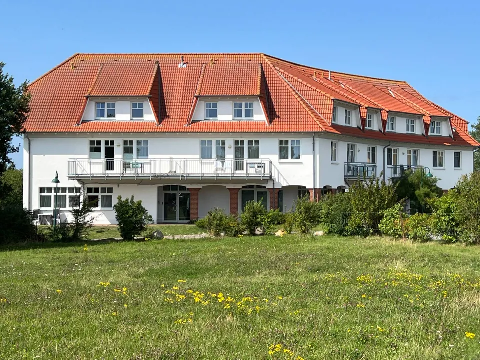 Ein langgestrecktes weißes Haus mit rotem Ziegeldach, Gauben und Balkonen, davor eine Wiese mit gelben Blüten.